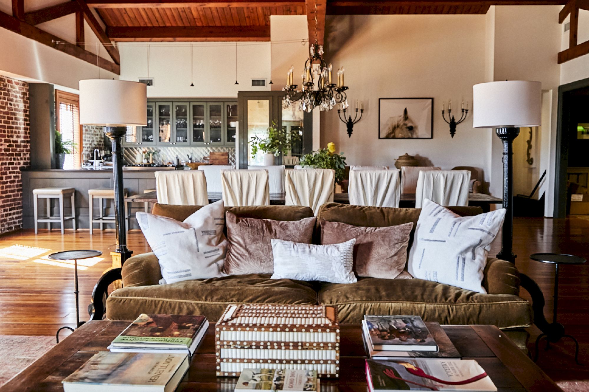 A cozy, stylish living area with a brown sofa and assorted throw pillows, a coffee table stacked with magazines, and a bright dining space in the background.