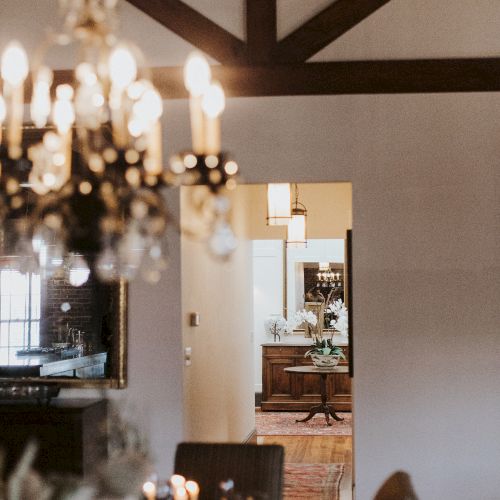 A cozy dining room with a chandelier, warm lighting, and a doorway opening to another seating area with a wooden table and decor, inviting ambiance.