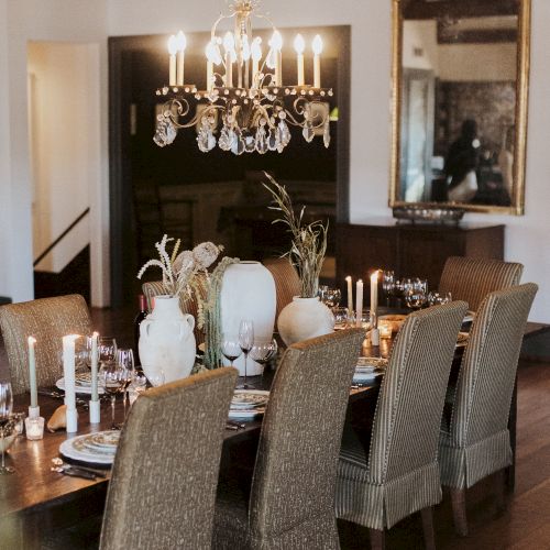 A long dining table with cream woven chairs, a crystal chandelier, white and neutral vases centerpieces, candles lit, and a warm, elegant room.
