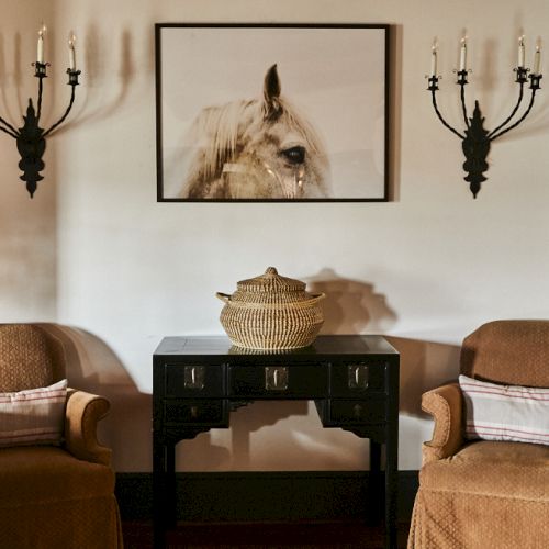 Two cushioned armchairs flank a black console table with a woven basket, framed horse portrait overhead, and candle sconces on either side.