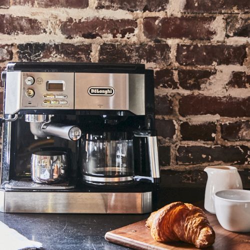 A stainless coffee machine sits on a bar counter with two cups and a croissant on a wooden board, against a brick wall backdrop.