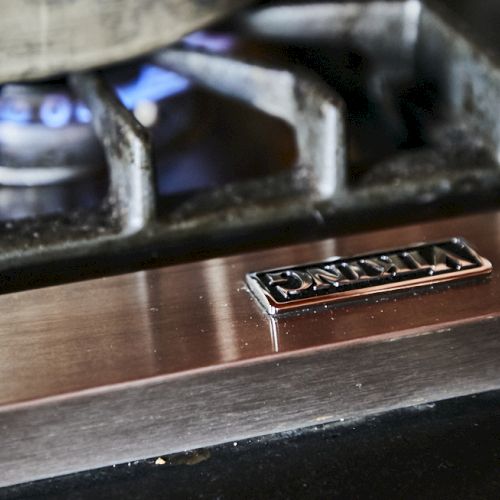 A close-up of a gas stove burner with a blue flame and a metal nameplate on a brushed metal surface, likely a vintage or stainless cooktop.