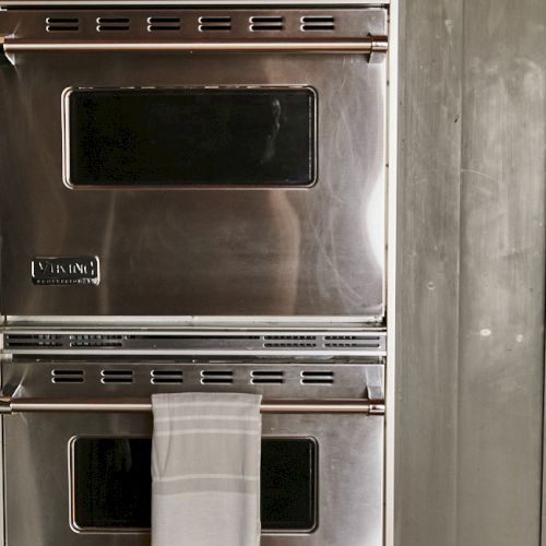 A vintage stainless steel double oven with a woven towel hanging from the lower door, built-in and slightly worn, in a muted kitchen corner.