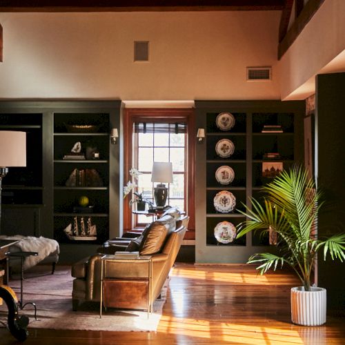 A cozy living room with dark built-in shelves, a tan leather chair, a floor lamp, warm wood floors, a large plant, and natural light streaming through a window, ending this sentence.