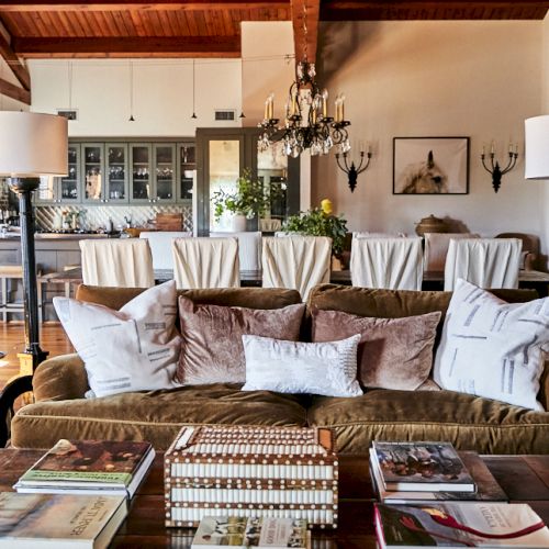 A stylish living room with a brown sofa and multiple cushions, a coffee table stacked with magazines, and a bright, open dining area in the background.