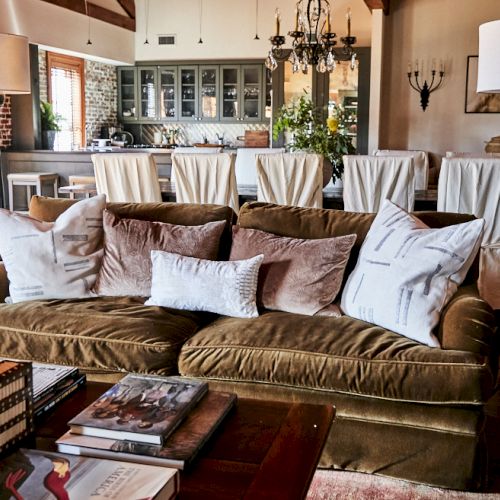 A cozy living room with a brown sofa, numerous white and beige pillows, wooden coffee table, chic lamps, and a bright dining area in the background.