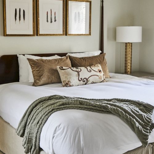 A cozy hotel-style bedroom with a large bed, white linens, decorative pillows, a throw blanket, and matching bedside tables with lamps.