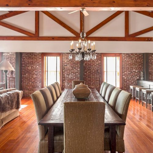 A spacious loft dining area with a long wooden table, upholstered chairs, brick walls, large windows, and a chandelier, plus a cozy living space at left.