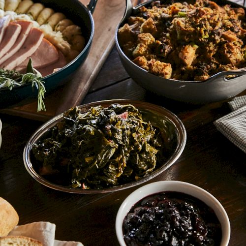 A rustic table with cooked meats, greens, and dark jams or chutneys; bread rolls and a cup of sauce complete a hearty spread.